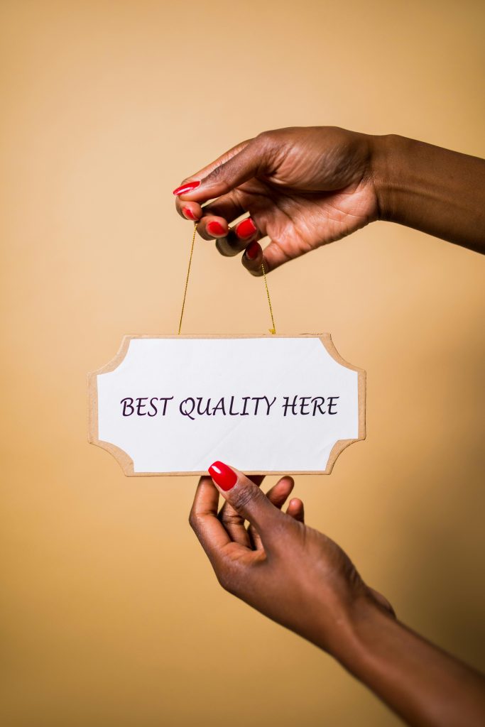 Stylish female hands with red nails holding a sign that says 'Best Quality Here' against a soft background.